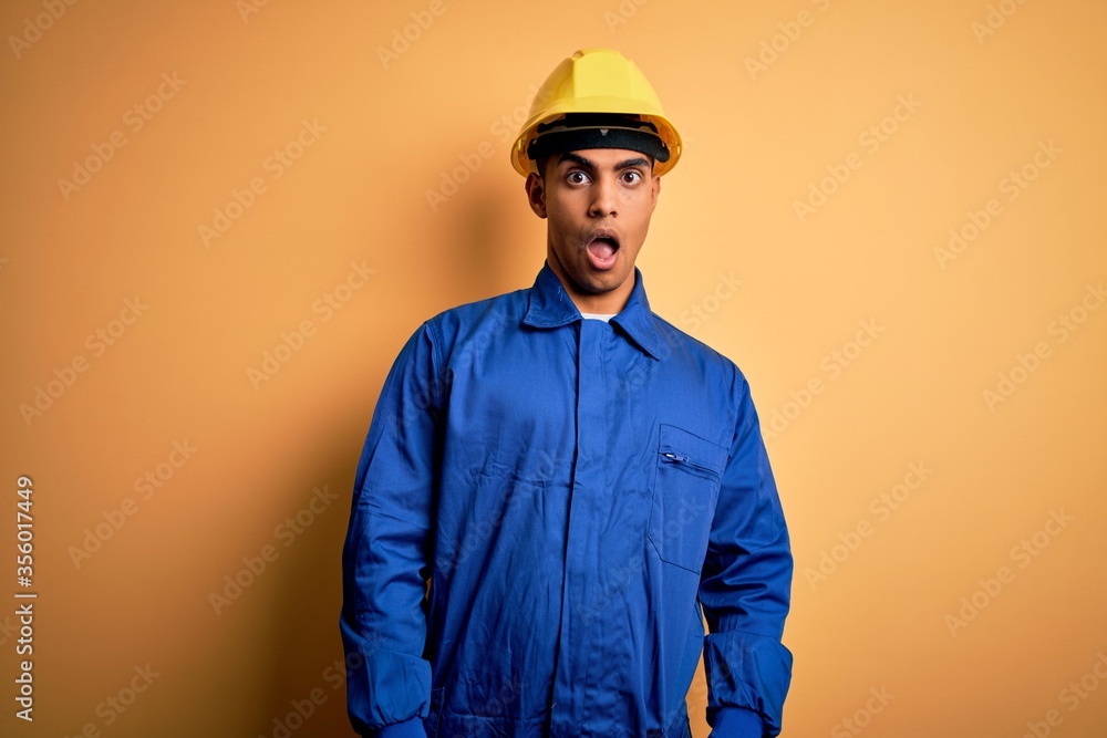 Young handsome african american worker man wearing blue uniform and security helmet In shock face, looking skeptical and sarcastic, surprised with open mouth