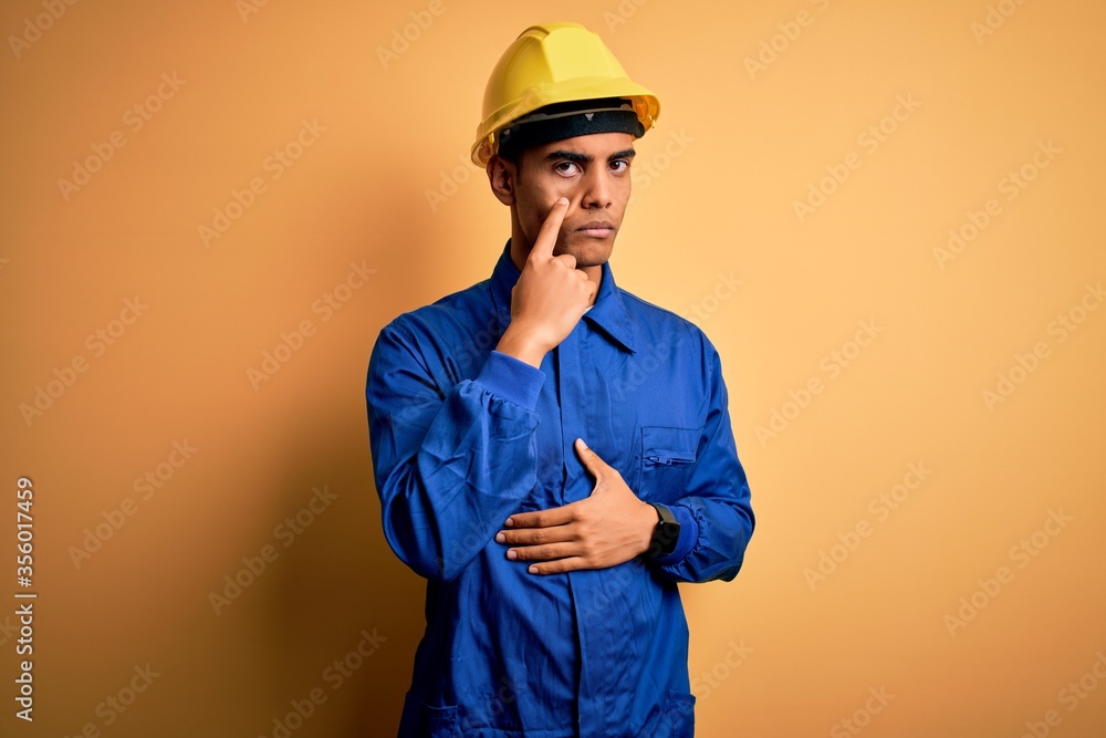 Young handsome african american worker man wearing blue uniform and security helmet Pointing to the eye watching you gesture, suspicious expression