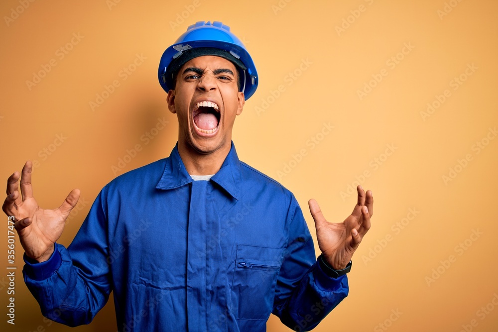 Young handsome african american worker man wearing blue uniform and security helmet crazy and mad shouting and yelling with aggressive expression and arms raised. Frustration concept.