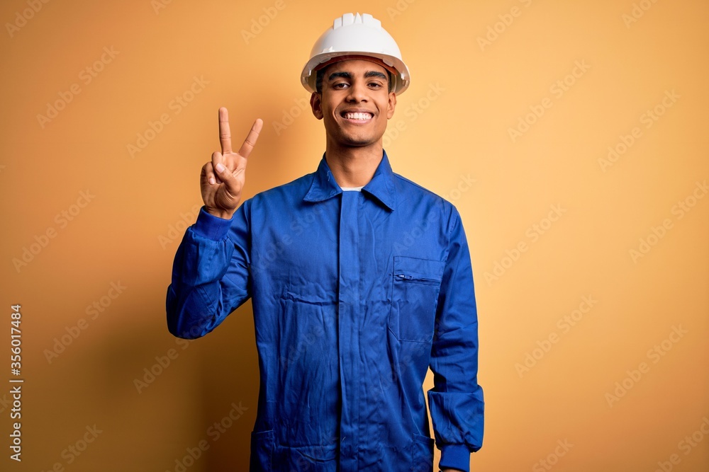 Young handsome african american worker man wearing blue uniform and security helmet smiling with happy face winking at the camera doing victory sign. Number two.