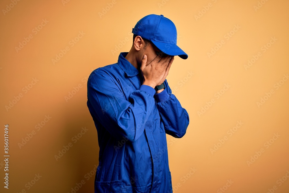 Young african american mechanic man wearing blue uniform and cap over yellow background with sad expression covering face with hands while crying. Depression concept.
