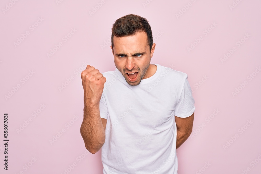 Handsome man with blue eyes wearing casual white t-shirt standing over pink background angry and mad raising fist frustrated and furious while shouting with anger. Rage and aggressive concept. Handsome man with blue eyes wearing casual white t-shirt standing over pink background angry and mad raising fist frustrated and furious while shouting with anger. Rage and aggressive concept.