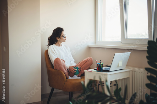 Thoughtful caucasian business girl with curly hair and eyeglasses drinking a cup of tea in front of pc near the window