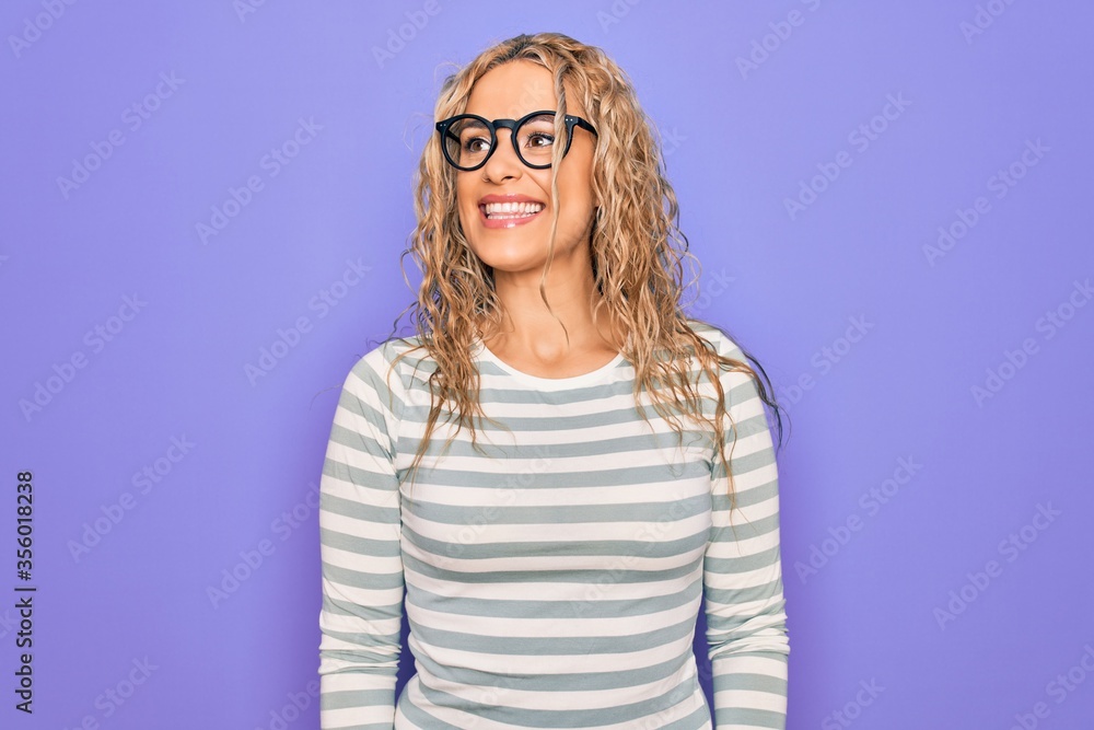 Beautiful blonde woman wearing casual striped t-shirt and glasses over purple background looking away to side with smile on face, natural expression. Laughing confident. Beautiful blonde woman wearing casual striped t-shirt and glasses over purple background looking away to side with smile on face, natural expression. Laughing confident.