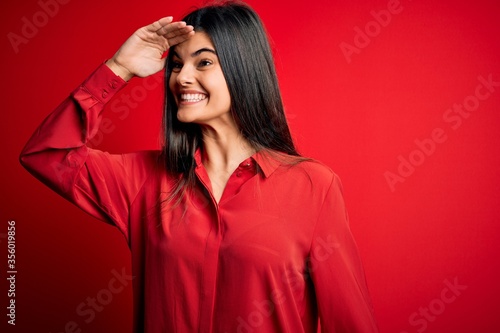 Young beautiful brunette woman wearing casual shirt standing over red background very happy and smiling looking far away with hand over head. Searching concept.