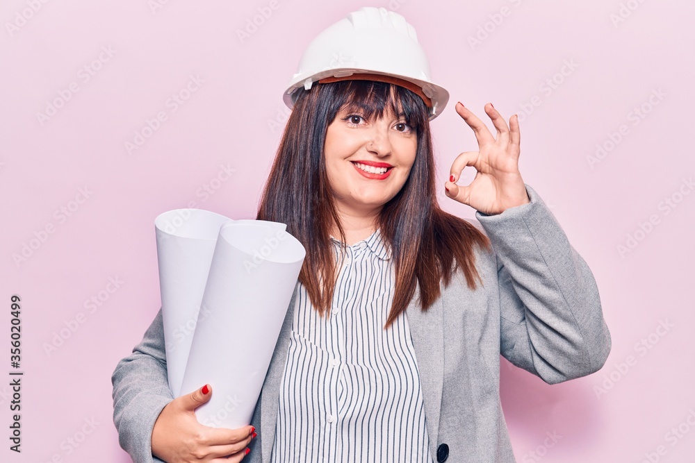 Young plus size woman holding paper blueprints doing ok sign with fingers, smiling friendly gesturing excellent symbol Young plus size woman holding paper blueprints doing ok sign with fingers, smiling friendly gesturing excellent symbol