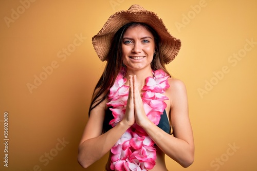 Young beautiful woman with blue eyes on vacation wearing bikini and hawaiian lei praying with hands together asking for forgiveness smiling confident.