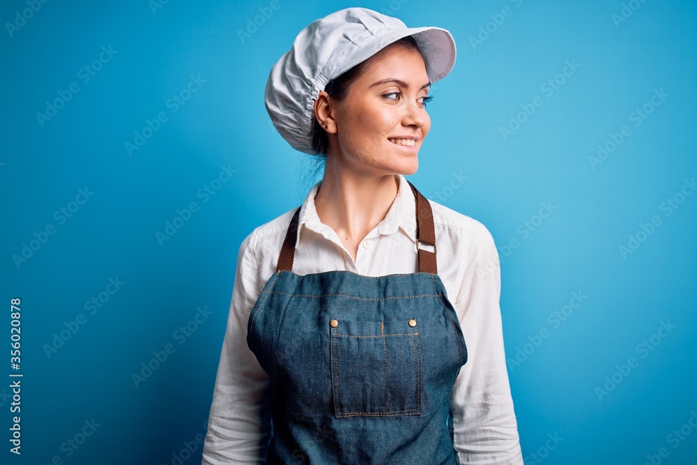 Young beautiful baker woman with blue eyes wearing apron and cap over blue background looking away to side with smile on face, natural expression. Laughing confident. Young beautiful baker woman with blue eyes wearing apron and cap over blue background looking away to side with smile on face, natural expression. Laughing confident.