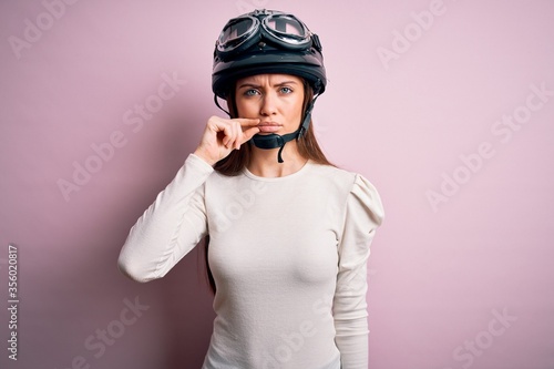 Young beautiful motorcyclist woman with blue eyes wearing moto helmet over pink background mouth and lips shut as zip with fingers. Secret and silent, taboo talking