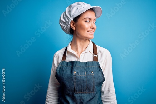 Young beautiful baker woman with blue eyes wearing apron and cap over blue background looking away to side with smile on face, natural expression. Laughing confident.