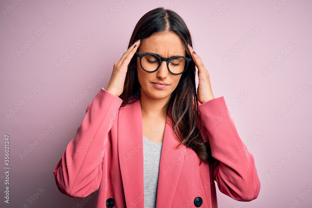 Young beautiful brunette businesswoman wearing jacket and glasses over pink background suffering from headache desperate and stressed because pain and migraine. Hands on head.