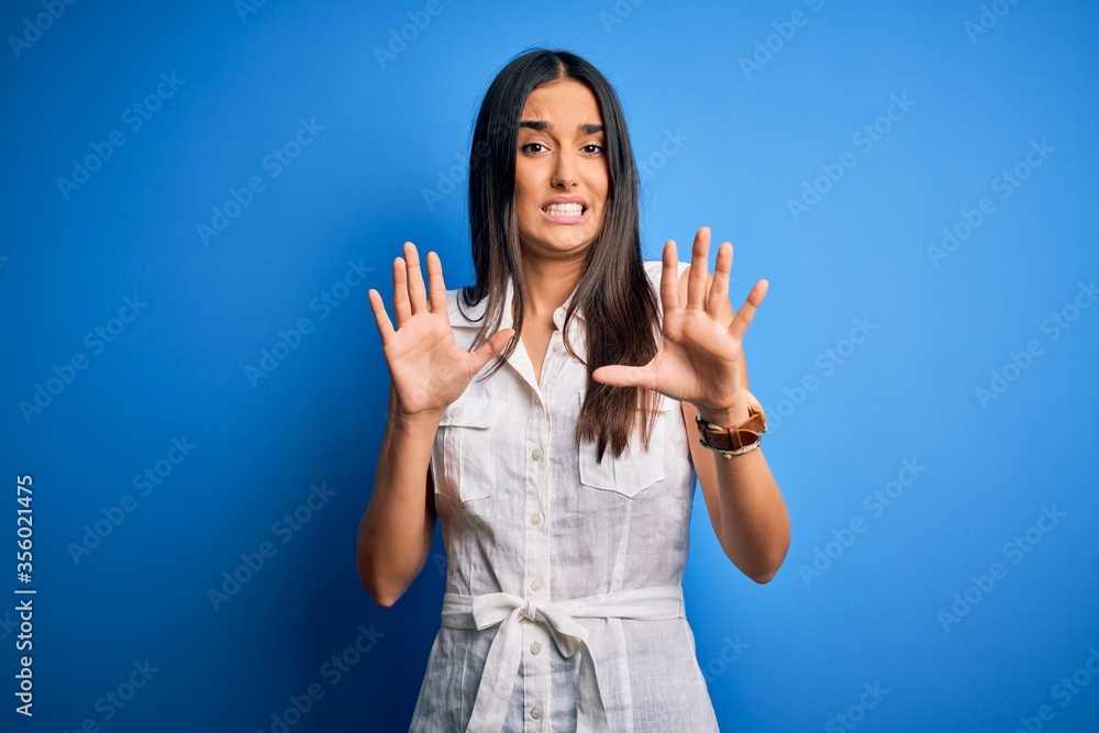 Young beautiful brunette woman wearing casual dress over isolated blue background afraid and terrified with fear expression stop gesture with hands, shouting in shock. Panic concept. Young beautiful brunette woman wearing casual dress over isolated blue background afraid and terrified with fear expression stop gesture with hands, shouting in shock. Panic concept.