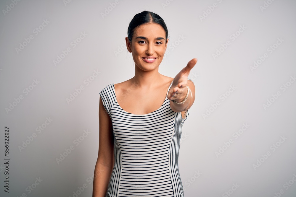 Young beautiful brunette woman wearing casual striped dress over isolated white background smiling friendly offering handshake as greeting and welcoming. Successful business. Young beautiful brunette woman wearing casual striped dress over isolated white background smiling friendly offering handshake as greeting and welcoming. Successful business.