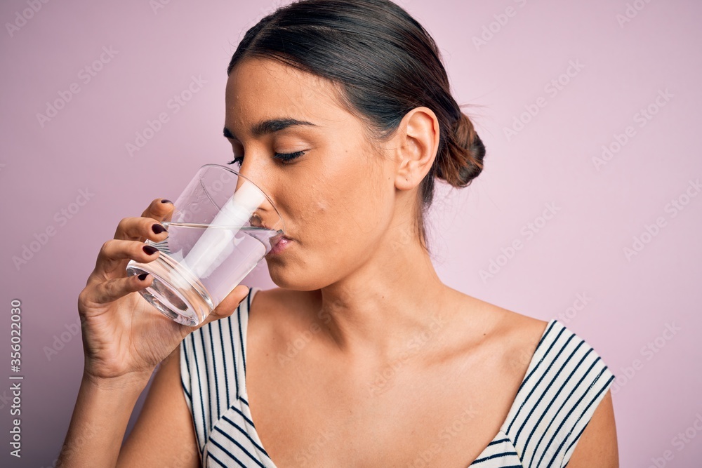 Young beautiful brunette woman drinking glass of healthy water to refreshment standing over isolated pink background Young beautiful brunette woman drinking glass of healthy water to refreshment standing over isolated pink background