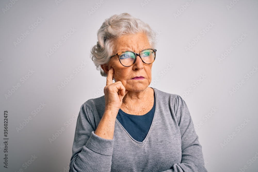 Senior beautiful grey-haired woman wearing casual sweater and glasses over white background with hand on chin thinking about question, pensive expression. Smiling with thoughtful face. Doubt concept. Senior beautiful grey-haired woman wearing casual sweater and glasses over white background with hand on chin thinking about question, pensive expression. Smiling with thoughtful face. Doubt concept.