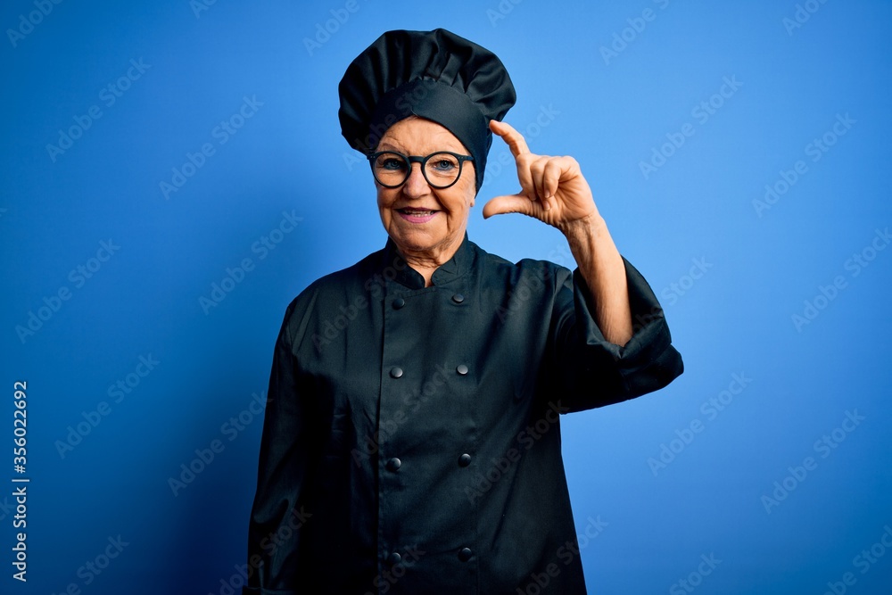 Senior beautiful grey-haired chef woman wearing cooker uniform and hat over blue background smiling and confident gesturing with hand doing small size sign with fingers looking and the camera. Measure Senior beautiful grey-haired chef woman wearing cooker uniform and hat over blue background smiling and confident gesturing with hand doing small size sign with fingers looking and the camera. Measure