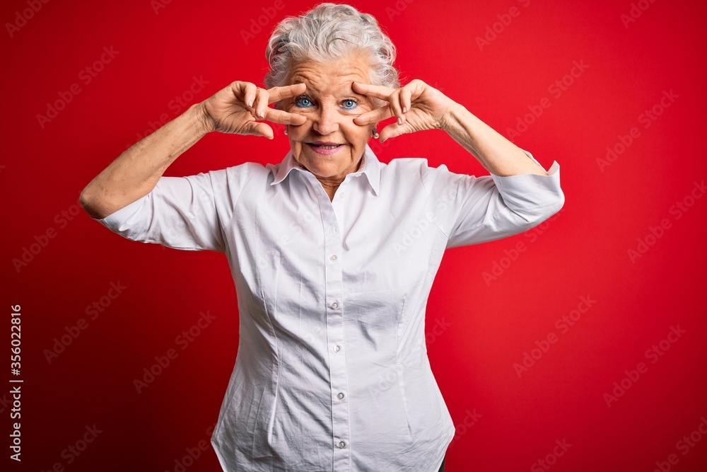 Senior beautiful woman wearing elegant shirt standing over isolated red background Doing peace symbol with fingers over face, smiling cheerful showing victory