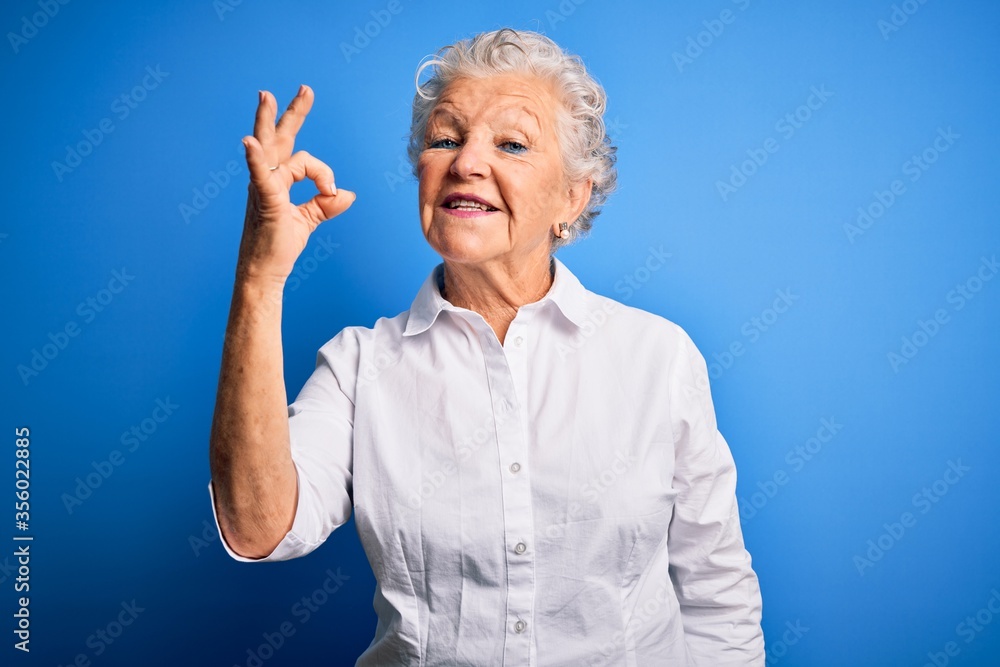 Senior beautiful woman wearing elegant shirt standing over isolated blue background smiling positive doing ok sign with hand and fingers. Successful expression. Senior beautiful woman wearing elegant shirt standing over isolated blue background smiling positive doing ok sign with hand and fingers. Successful expression.