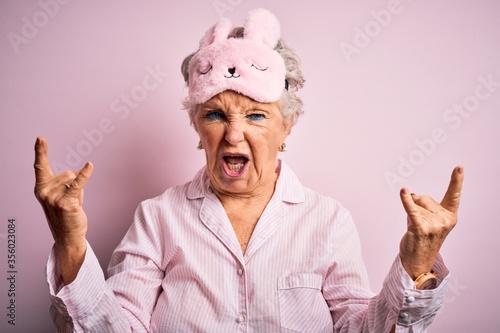 Canvastavla Senior beautiful woman wearing sleep mask and pajama over isolated pink background shouting with crazy expression doing rock symbol with hands up