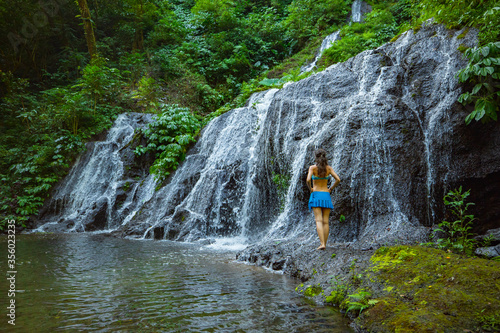 Young traveler woman at waterfall in tropical forest. View from back. Travel concept. Pucak Manik waterfall Wanagiri, Bali. Slow shutter speed, motion photography.