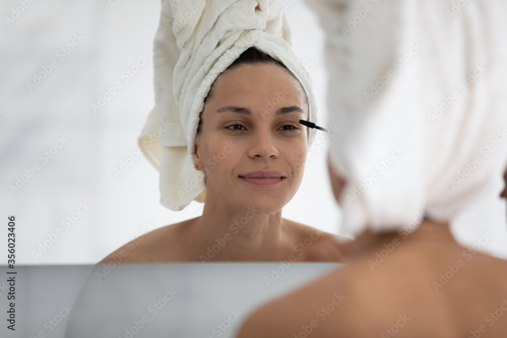 Head shot close up beautiful young woman applying black mascara on eyelashes, attractive girl wearing white towel on head putting daily makeup, looking in mirror, using cosmetics product Head shot close up beautiful young woman applying black mascara on eyelashes, attractive girl wearing white towel on head putting daily makeup, looking in mirror, using cosmetics product