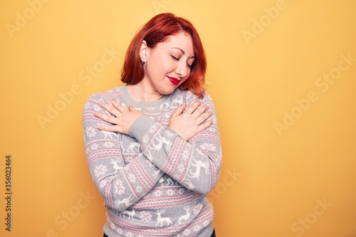 Beautiful redhead woman wearing christmas sweater with reindeer over yellow background hugging oneself happy and positive, smiling confident. Self love and self care © Krakenimages.com