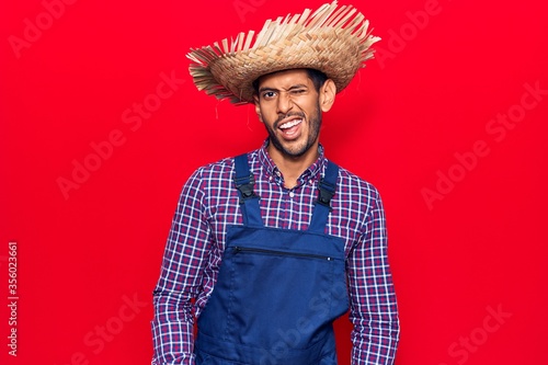 Young latin man wearing farmer hat and apron winking looking at the camera with sexy expression, cheerful and happy face.