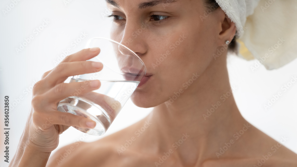 Close up beautiful woman wearing bath towel on head drinking pure still mineral water, holding glass in hand, thirsty girl enjoying clean water, healthy lifestyle and habit concept, natural beauty Close up beautiful woman wearing bath towel on head drinking pure still mineral water, holding glass in hand, thirsty girl enjoying clean water, healthy lifestyle and habit concept, natural beauty