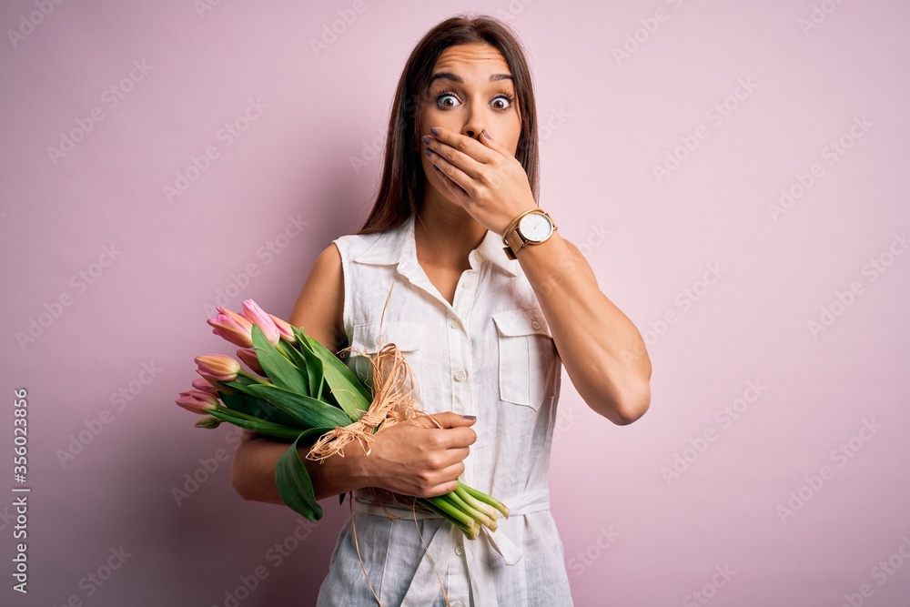 Young beautiful brunette woman holding bouquet of tulips flowers over pink background shocked covering mouth with hands for mistake. Secret concept. Young beautiful brunette woman holding bouquet of tulips flowers over pink background shocked covering mouth with hands for mistake. Secret concept.