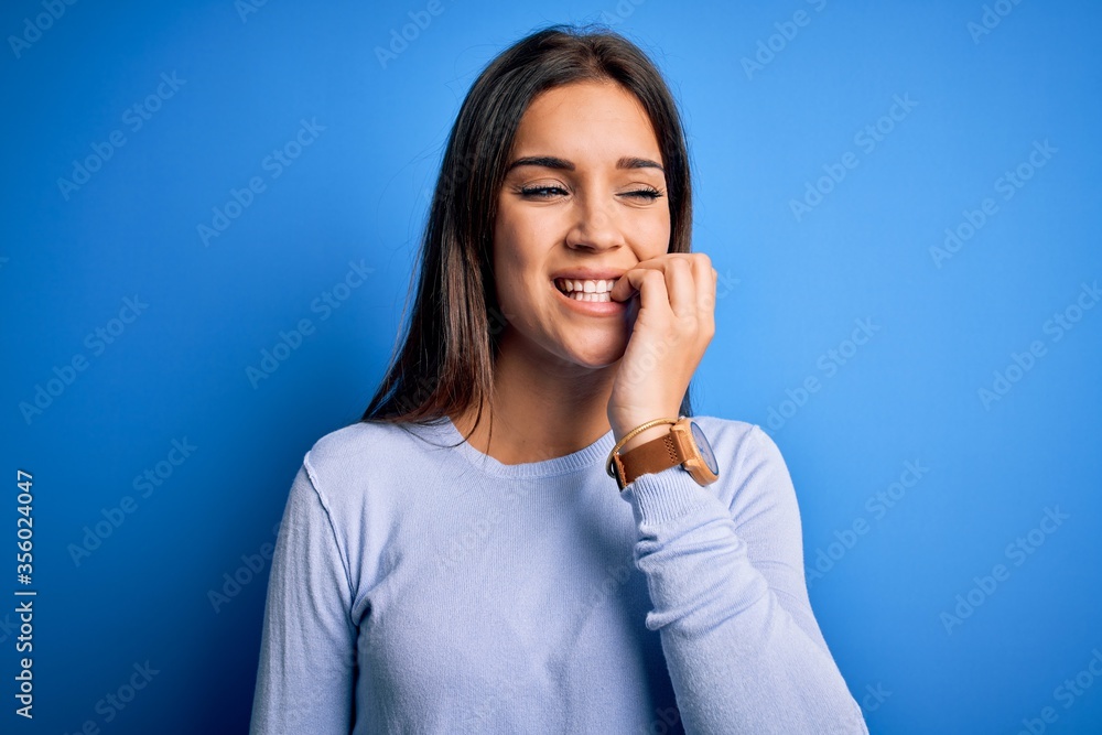 Fototapeta premium Young beautiful brunette woman wearing casual sweater standing over blue background looking stressed and nervous with hands on mouth biting nails. Anxiety problem.