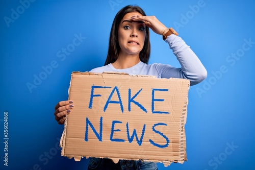 Young beautiful brunette woman holding banner with fake news message stressed with hand on head, shocked with shame and surprise face, angry and frustrated. Fear and upset for mistake. © Krakenimages.com