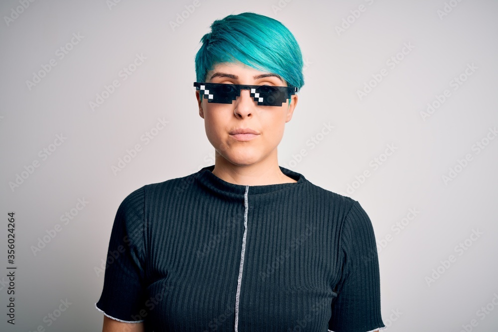 Young woman with blue fashion hair wearing thug life sunglasses over white background with serious expression on face. Simple and natural looking at the camera. Young woman with blue fashion hair wearing thug life sunglasses over white background with serious expression on face. Simple and natural looking at the camera.
