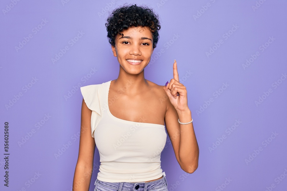 Young beautiful african american afro woman wearing casual t-shirt over purple background showing and pointing up with finger number one while smiling confident and happy. Young beautiful african american afro woman wearing casual t-shirt over purple background showing and pointing up with finger number one while smiling confident and happy.