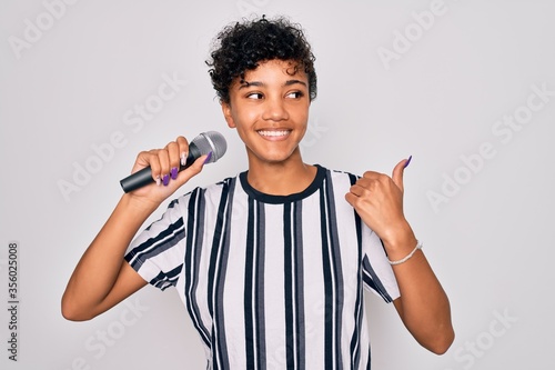 Beautiful african american afro singer woman singing using microphone over white background pointing and showing with thumb up to the side with happy face smiling