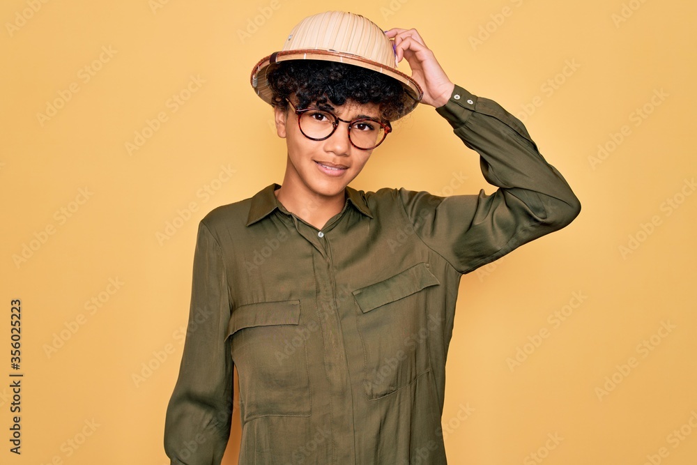 Young beautiful african american tourist woman wearing glasses and explorer hat confuse and wonder about question. Uncertain with doubt, thinking with hand on head. Pensive concept. Young beautiful african american tourist woman wearing glasses and explorer hat confuse and wonder about question. Uncertain with doubt, thinking with hand on head. Pensive concept.
