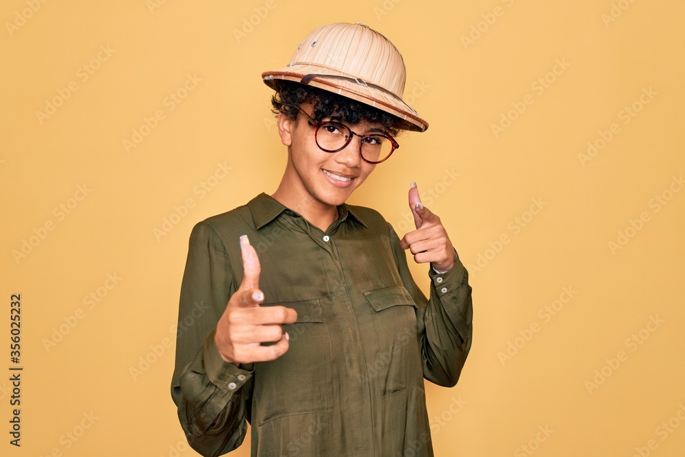 Young beautiful african american tourist woman wearing glasses and explorer hat pointing fingers to camera with happy and funny face. Good energy and vibes. Young beautiful african american tourist woman wearing glasses and explorer hat pointing fingers to camera with happy and funny face. Good energy and vibes.