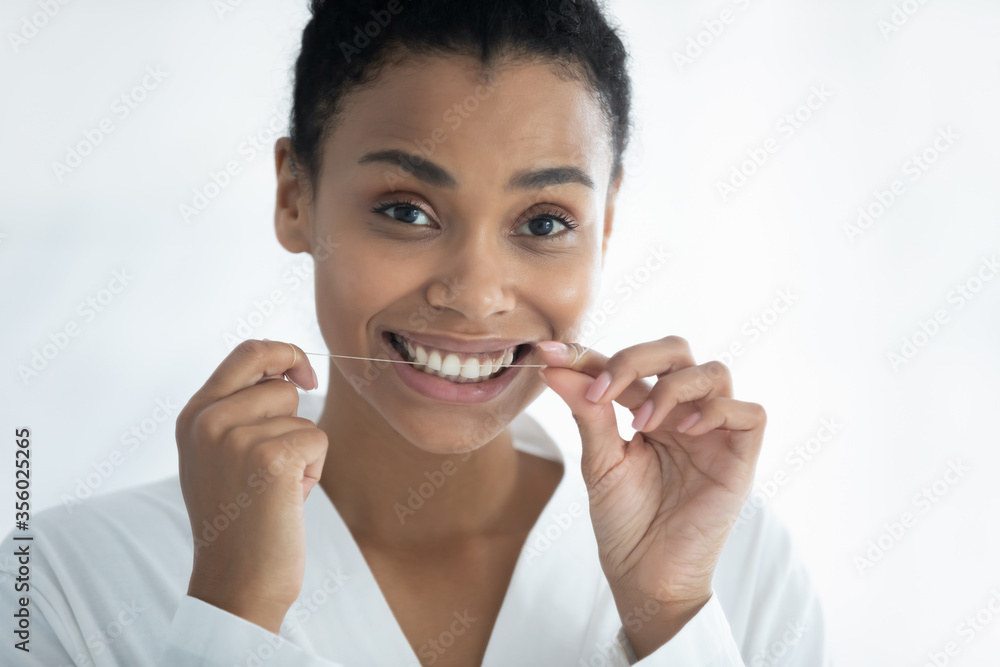 Head shot portrait smiling African American young woman holding dental floss, looking at camera, happy beautiful girl wearing white bathrobe removing food cleaning teeth, oral hygiene concept Head shot portrait smiling African American young woman holding dental floss, looking at camera, happy beautiful girl wearing white bathrobe removing food cleaning teeth, oral hygiene concept