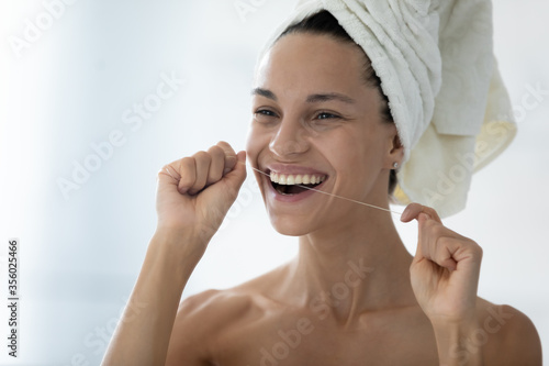 Head shot smiling beautiful woman holding using dental floss, removing food cleaning teeth, standing in bathroom with white bath towel on head, healthcare and personal hygiene, morning routine