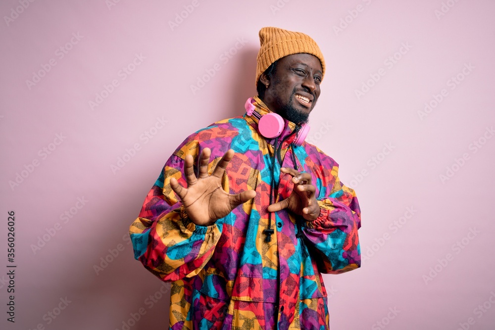 Young handsome african american man wearing colorful coat and cap over pink background disgusted expression, displeased and fearful doing disgust face because aversion reaction. With hands raised