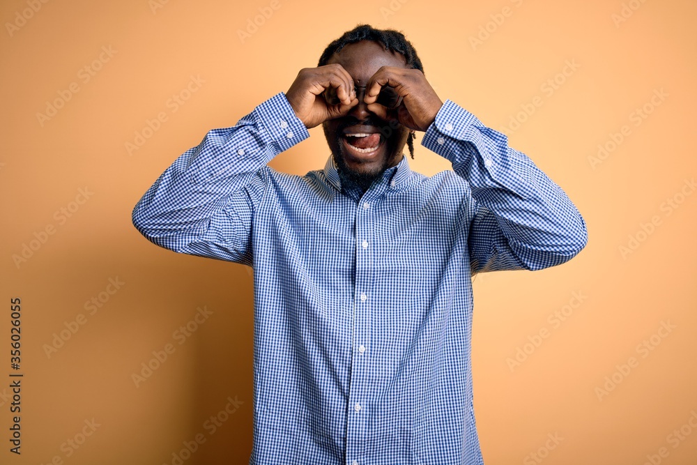 Young handsome african american man wearing shirt and glasses over yellow background doing ok gesture like binoculars sticking tongue out, eyes looking through fingers. Crazy expression. Young handsome african american man wearing shirt and glasses over yellow background doing ok gesture like binoculars sticking tongue out, eyes looking through fingers. Crazy expression.