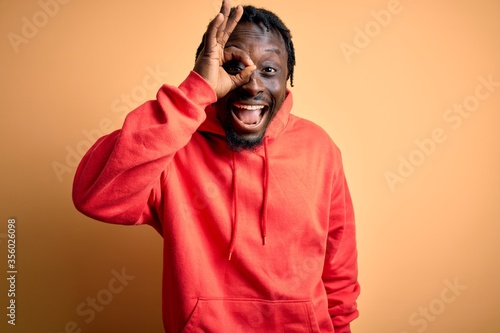 Young african american sporty man wearing sweatshirt with hoodie over yellow background doing ok gesture with hand smiling, eye looking through fingers with happy face.