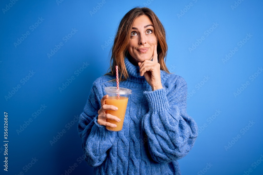 Young beautiful brunette woman drinking healthy orange juice over blue background serious face thinking about question, very confused idea Young beautiful brunette woman drinking healthy orange juice over blue background serious face thinking about question, very confused idea