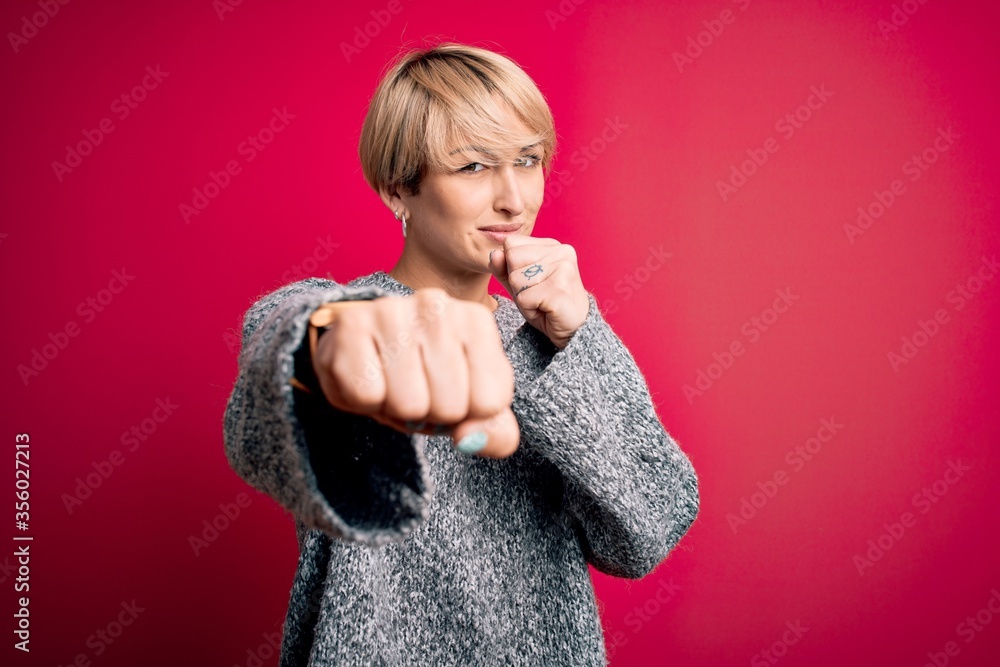 Young blonde woman with modern short hair wearing casual sweater over pink background Punching fist to fight, aggressive and angry attack, threat and violence Young blonde woman with modern short hair wearing casual sweater over pink background Punching fist to fight, aggressive and angry attack, threat and violence