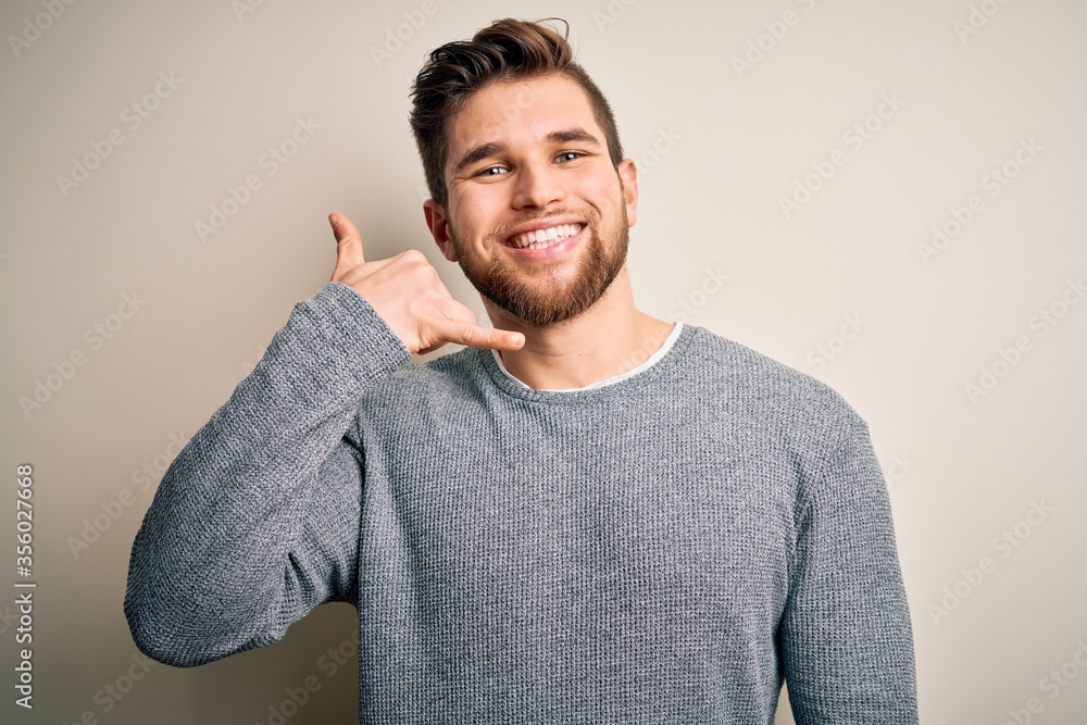 Young handsome blond man with beard and blue eyes wearing casual sweater smiling doing phone gesture with hand and fingers like talking on the telephone. Communicating concepts. Young handsome blond man with beard and blue eyes wearing casual sweater smiling doing phone gesture with hand and fingers like talking on the telephone. Communicating concepts.