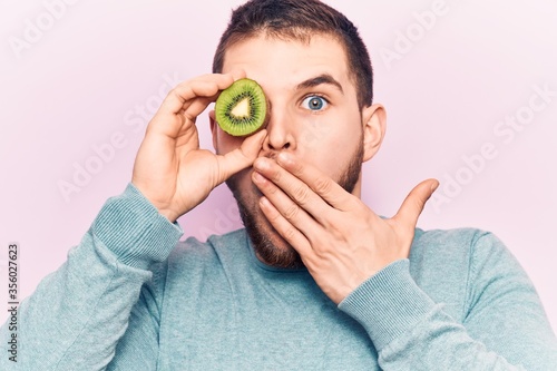 Young handsome man holding slice of kiwi over eye covering mouth with hand, shocked and afraid for mistake. surprised expression