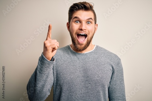 Young handsome blond man with beard and blue eyes wearing casual sweater pointing finger up with successful idea. Exited and happy. Number one.