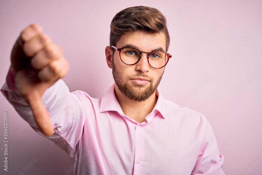 Young handsome blond man with beard and blue eyes wearing pink shirt and glasses looking unhappy and angry showing rejection and negative with thumbs down gesture. Bad expression. Young handsome blond man with beard and blue eyes wearing pink shirt and glasses looking unhappy and angry showing rejection and negative with thumbs down gesture. Bad expression.