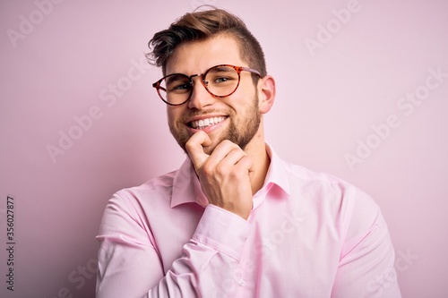 Young handsome blond man with beard and blue eyes wearing pink shirt and glasses looking confident at the camera with smile with crossed arms and hand raised on chin. Thinking positive.