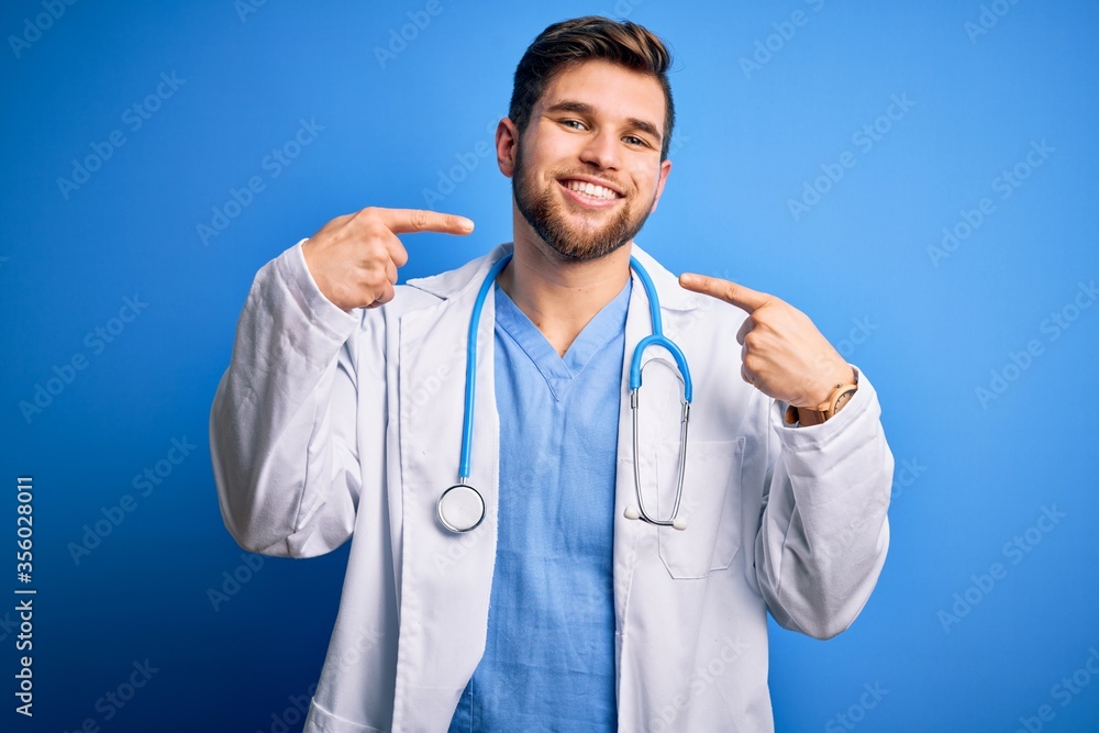 Young blond doctor man with beard and blue eyes wearing white coat and stethoscope smiling cheerful showing and pointing with fingers teeth and mouth. Dental health concept. Young blond doctor man with beard and blue eyes wearing white coat and stethoscope smiling cheerful showing and pointing with fingers teeth and mouth. Dental health concept.