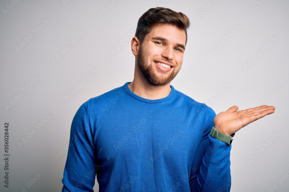 Young handsome blond man with beard and blue eyes wearing casual sweater smiling cheerful presenting and pointing with palm of hand looking at the camera. Young handsome blond man with beard and blue eyes wearing casual sweater smiling cheerful presenting and pointing with palm of hand looking at the camera.